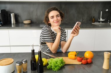 Modern genç bir kadının portresi ne pişireceğini düşünüyor, akıllı telefon ve sebzelerle mutfakta oturuyor, yemek yapıyor, sağlıklı diyet salatası yapıyor..