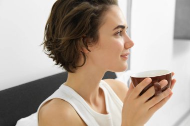 Şehvetli sabah konsepti. Genç, güzel bir kadın yatakta kahve içiyor, elinde kupa tutuyor ve taze demlenmiş içeceğin güzel kokusunu alıyor..