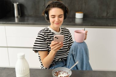 Çay içen ve kulaklıkla müzik dinleyen genç bir kadının portresi, mutfakta yemek yerken sosyal medyayı kaydırmak, mutlu bir şekilde gülümsemek..