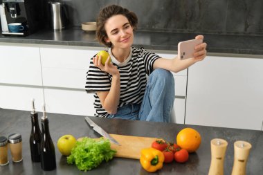 Mutfakta gülümseyen esmer kadının portresi, elmayla poz vermek, akıllı telefondan selfie çekmek, yemek pişirmek ve internet günlüğü için resim yapmak..