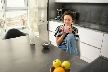 Şirin esmer kadın, sabah kahvaltı yapan öğrenci, sıcak çay içiyor, gülümsüyor ve memnun görünüyor..