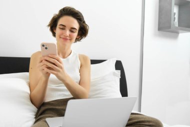 Modern bir kadın tasviri, yatakta dizüstü bilgisayarla ve akıllı telefonla dinleniyor, mesaj gönderiyor, cep telefonundan bildirimi okuyor..