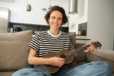 Tutkulu genç bir kadın, müzisyen evde ukulele çalıyor, neşeyle şarkı söylüyor, oturma odasında kanepede oturuyor..