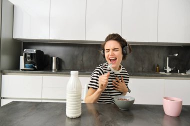 Mutlu ve duygusal genç kadın, kahvaltı yaparken şarkı söylüyor, mısır gevreğini sütle yiyor, kablosuz kulaklıkla müzik dinliyor, mutfakta oturuyor..
