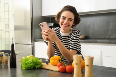 Güzel gülümseyen bir kadının portresi, evde yemek pişirmek, akıllı telefona bakmak, sağlıklı vejetaryen tarifi aramak, sebzelerin yanında durmak, kendine salata yapmak..