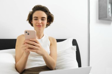 Modern bir kadın tasviri, yatakta dizüstü bilgisayarla ve akıllı telefonla dinleniyor, mesaj gönderiyor, cep telefonundan bildirimi okuyor..