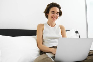 Modern kadın evde dinleniyor, dizüstü bilgisayarla yatakta oturuyor, bilgisayar ekranına bakıyor, gülüyor ve gülümsüyor..