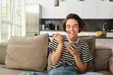 Modern kadın portresi, mısır gevreğini sütle yiyen ve kanepede oturan, TV karşısında dinlenen, evde kahvaltı yapan.