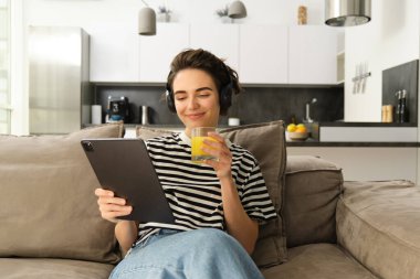 Kadın portresi evde oturuyor, dijital tabletle kablosuz kulaklık takıyor, taze portakal suyu içiyor..