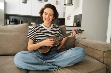 Genç, mutlu bir kadın kanepede oturup ukulele çalıyor, şarkı söylüyor ve yeni bir müzik aleti öğrenmekten zevk alıyor..