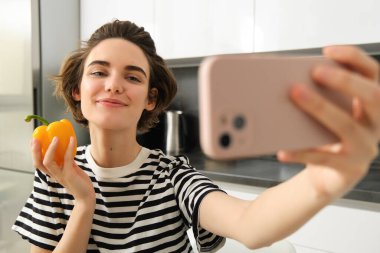 Gülümseyen, güzel genç bir kadın taze sarı biberle selfie çekiyor, yemek blogcusu mutfakta sebzelerle selfie çekiyor, vegan sosyal medya için yemek yapıyor ve video kaydediyor..