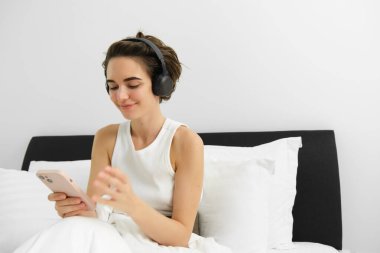 Yatağa giden genç bir kadının portresi, kulaklık takan, akıllı telefon kullanan, podcast ya da istiridye dinleyen, kulaklıkla müzik dinleyen..