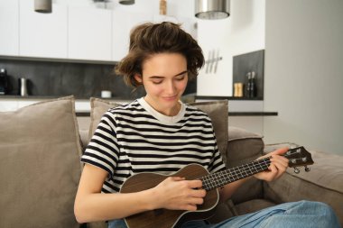 Ukulele çalan, şarkı söyleyen, gülen, evinde oturan neşeli genç bir kadının portresi. Yaşam tarzı ve müzik konsepti