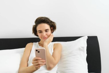 Yaşam tarzı ve teknoloji konsepti. Gülümseyen güzel bir kadın akıllı telefonuyla yatakta uzanıyor, yatak odasında dinleniyor, sosyal medyada sohbet ediyor, online alışveriş yapıyor..