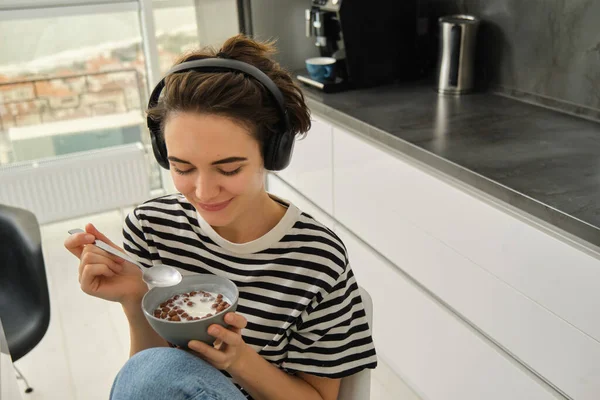 Close up portrait of smiling, beautiful young woman in headphones, eating cereals for breakfast and listening music or e-book, holding spoon with bowl.