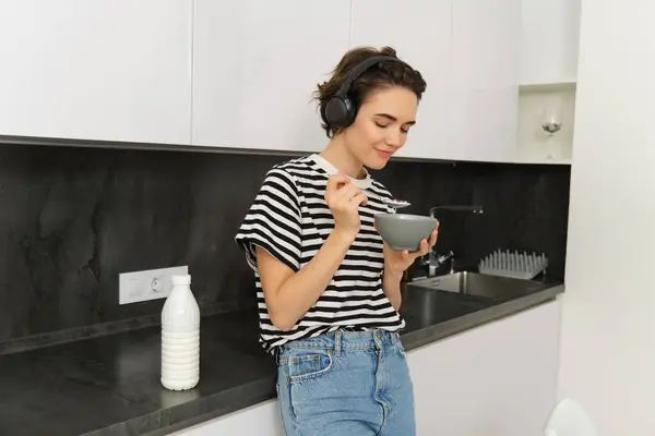 Candid young woman eats breakfast, cereals from bowl, stands near kitchen counter, listens music in wireless headphones.