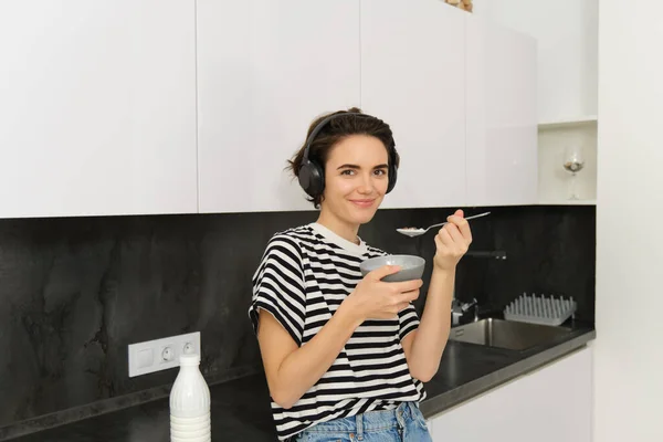 Cute young woman eats cereals with milk, holds spoon near mouth and smiles, listens music in wireless headphones, sits in kitchen.