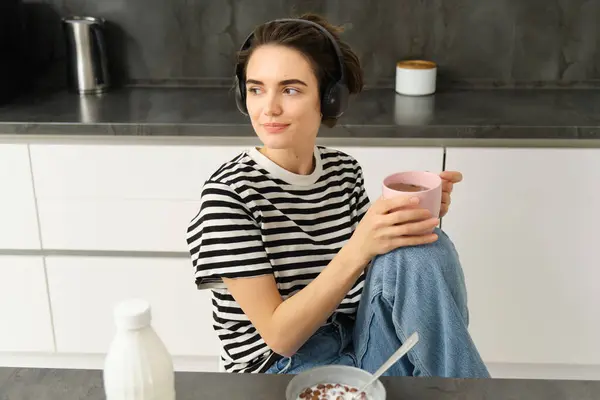 Portrait of stylish modern woman in headphones, listens music and drinks tea, eats cereals with milk for lunch, sits in kitchen, looks outside window with relaxed smile.