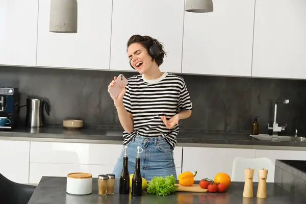 Carefree woman cooking in the kitchen, singing along favourite song, listening music in wireless headphones, holding mobile phone, chopping vegetables, preparing healthy meal.