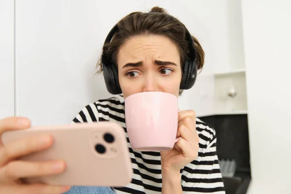 Portrait of woman with sad and worried face, watching intense tv series on smartphone app, drinking tea and staring at mobile phone screen.