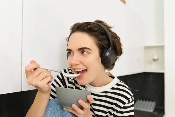 Happy beautiful woman, eating breakfast, cereals with milk as morning meal, holding bowl and spoon, listening music in wireless earphones, sitting in kitchen.