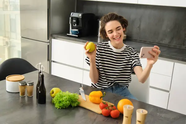 Smiling young woman, food blogger recording a video on smartphone with food, cooking and making selfie, taking pictures during meal preparation, sitting near vegetables and holding an apple.