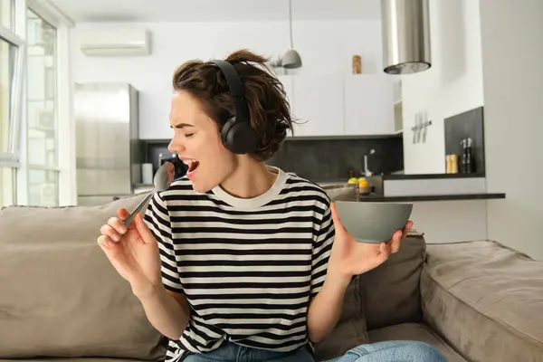 Close up portrait of excited woman in headphones, singing and listening music while eating cereals for breakfast, using spoon as a microphone, sitting in living room at home.