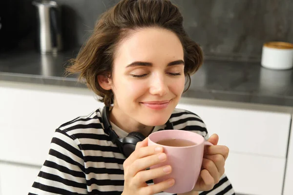 Close up portrait of woman with closed eyes and pleased smile, drinks warm tea with satisfaction, sitting in cosy kitchen.