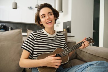 Güzel genç bir kadın ukulele çalıyor, şarkı söylüyor ve müzik aletinin tadını çıkarıyor, evde tek başına vakit geçiriyor, oturma odasında dinleniyor..