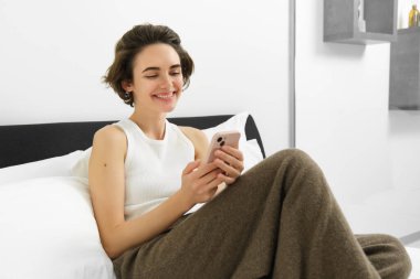 Genç güzel bir kadının portresi yatakta uzanıyor, yatak odasında dinleniyor, mesajlaşıyor, cep telefonu kullanıyor, akıllı telefonu tutuyor ve gülümsüyor, sosyal medyada kayıyor, evde rahat zaman geçiriyor..