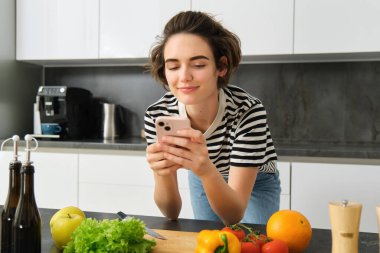 Genç gülümseyen bir kadının portresi, akıllı telefondan yemek tarifi araması, sebze ve kesim tahtası yanında durmak, yemek yapmak, salata hazırlamak ve cep telefonunda sosyal medya kullanmak..