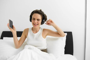 Yatakta mutlu genç bir kadının portresi, kablosuz kulaklıkla müzik dinlemek, yatak odasında dans etmek, cep telefonunu elinde tutmak. Boşluğu kopyala