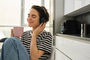 Yaşam tarzı ve müzik konsepti. Bir fincan çayla gülümseyen mutlu kadın, mutfakta oturur ve kulaklıklara dokunur, rahatlamış yüzüyle müzik dinler..