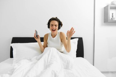 Yatağında şarkı söyleyen mutlu bir kadının portresi uyanıyor ve elinde cep telefonu olan kablosuz kulaklıkla müzik dinliyor..