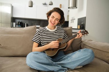 Ukulele çalan, şarkı söyleyen, gülen, evinde oturan neşeli genç bir kadının portresi. Yaşam tarzı ve müzik konsepti
