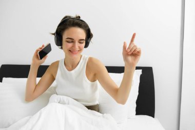 Yatağında dans eden, kablosuz kulaklıkla müzik dinleyen, cep telefonu tutan mutlu gülümseyen kaygısız bir kadın. Boşluğu kopyala