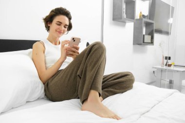 Akıllı telefon kullanan, yatakta oturan, yatak odasında dinlenen, sosyal medyada veya cep telefonunda web sitelerinde gezinen güzel kadın modelin portresi.