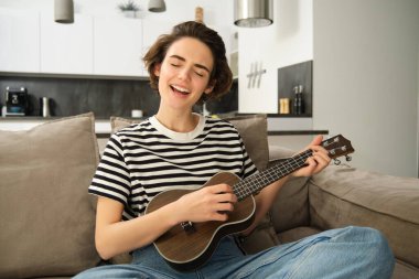 Güzel genç bir kadın ukulele çalıyor, şarkı söylüyor ve müzik aletinin tadını çıkarıyor, evde tek başına vakit geçiriyor, oturma odasında dinleniyor..