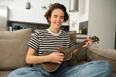 Kaygısız genç bayan model, kahverengi konser ukulele çalıyor, tutkuyla şarkı söylüyor, elinde küçük gitarla kanepede oturuyor..