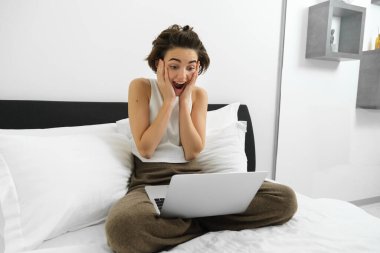 Evdeki kadın görüntüsü, yatakta oturmuş dizüstü bilgisayara bakıyor, şaşırmış, mutlu yüz ifadesiyle ekrana bakıyor..