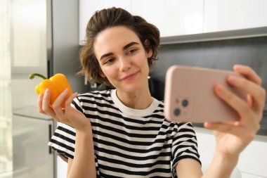 Gülümseyen, güzel genç bir kadın taze sarı biberle selfie çekiyor, yemek blogcusu mutfakta sebzelerle selfie çekiyor, vegan sosyal medya için yemek yapıyor ve video kaydediyor..