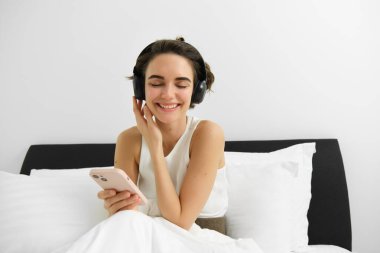 Yatakta güzel bir kız, kablosuz kulaklıkla müzik dinliyor, akıllı telefon kullanıyor ve gülümsüyor. Boşluğu kopyala