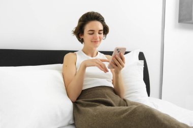 Yatak odasında güzel bir kadın modelin portresi, telefonu tutmak, akıllı telefona mesaj yazmak, yatakta dinlenmek ve gülümsemek..