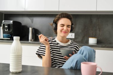 Güzel genç bir bayan model, mutfakta kahvaltı yapıyor, mısır gevreğini sütlü yiyor, gülümsüyor ve kahve içiyor, müzik dinliyor ya da kablosuz kulaklıkla podcast yapıyor..