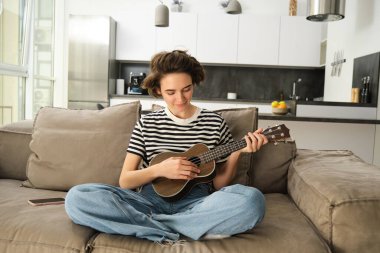 Kanepede oturan sevimli genç bir kadının portresi, gitar çalmayı öğreniyor, müzik aletini tutuyor, akor seçiyor, oturma odasında dinleniyor..