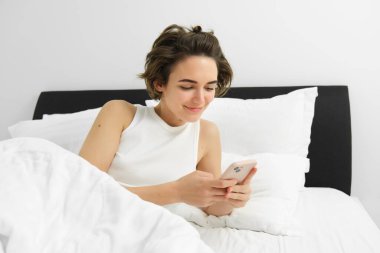 Genç bir kadının portresi yatakta uyanır, akıllı telefon kullanır, cep telefonunun alarmını kapatır, yatak odasında beyaz çarşafların üzerinde yatar..