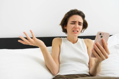 Kafası karışmış bir kadının portresi. Akıllı telefona bakıyor, omuz silkiyor, mobil ekrana bakıyor. Suratı asık..