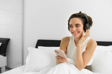 Yatakta güzel bir kız, kablosuz kulaklıkla müzik dinliyor, akıllı telefon kullanıyor ve gülümsüyor. Boşluğu kopyala