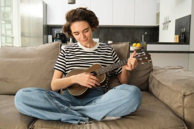 Yaşam tarzı ve hobi konsepti. Kanepede gülümseyen genç bir kadın, ukulele çalıyor, en sevdiği şarkı için tıngırdatıyor ve öğreniyor..