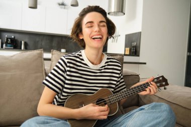 Genç, mutlu bir kadın kanepede oturup ukulele çalıyor, şarkı söylüyor ve yeni bir müzik aleti öğrenmekten zevk alıyor..